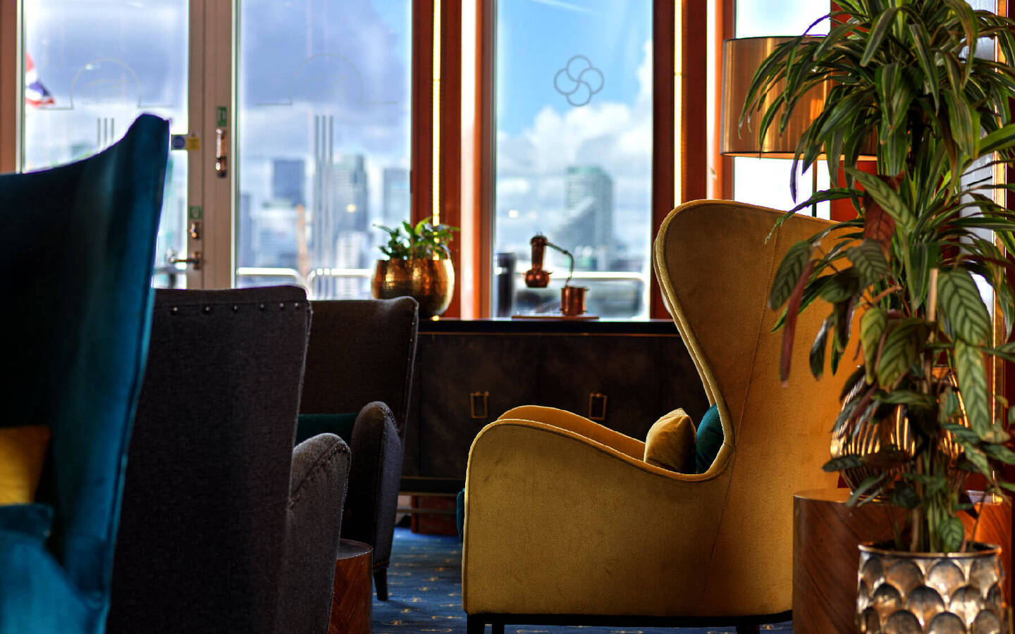 Cozy hotel lounge with velvet armchairs, plants and warm lamp by large windows showing a blurred city skyline