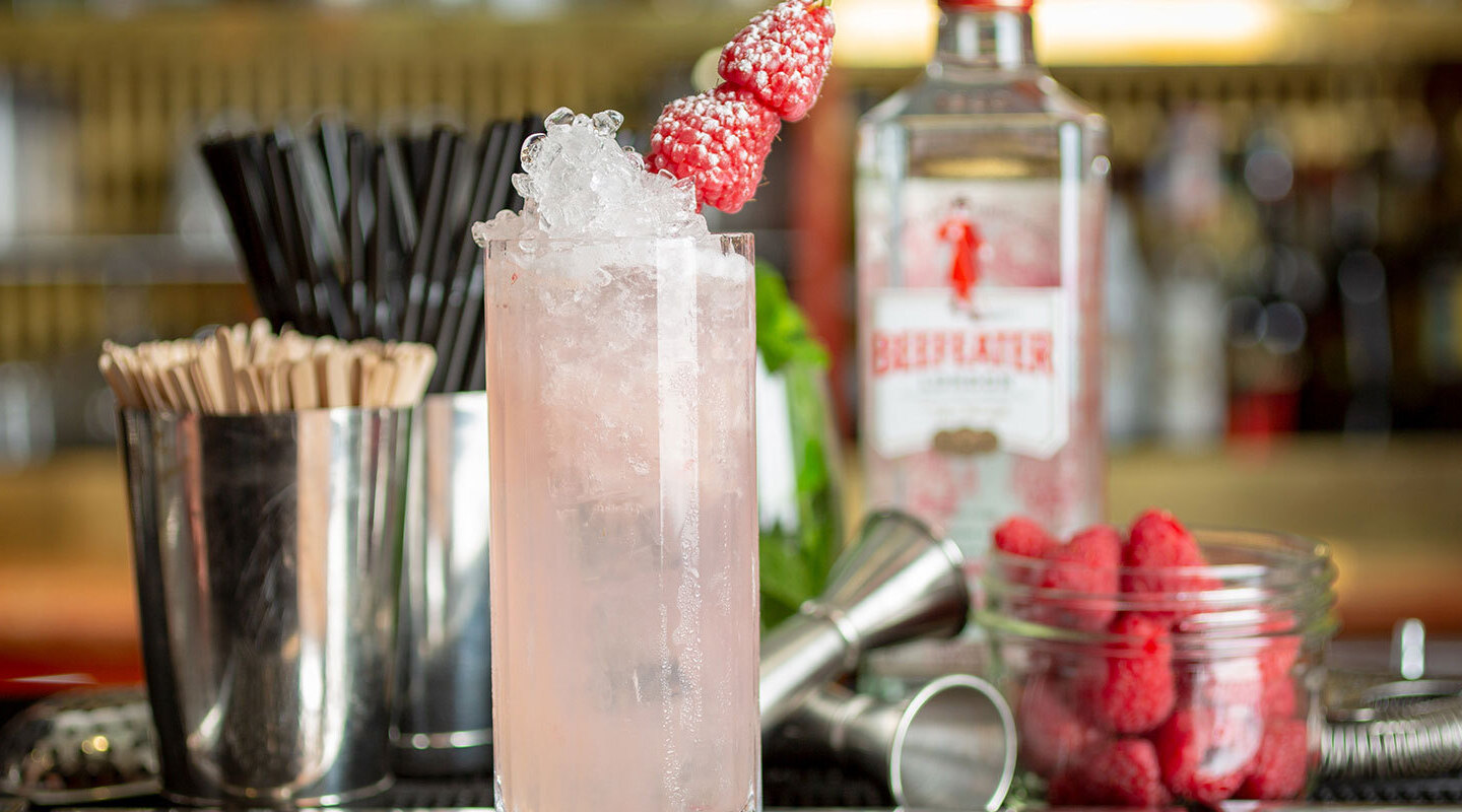 Pink gin cocktail with crushed ice topped by skewered raspberries at a hotel bar, surrounded by fresh raspberries and bartending tools