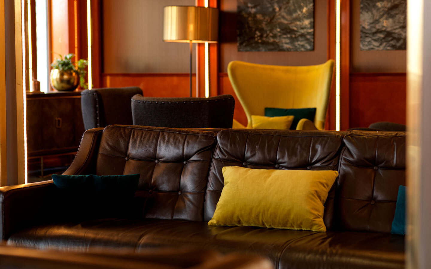 Cozy hotel lounge with tufted leather sofa, yellow accent pillows, velvet wing chair, warm wood panels and ambient lighting.