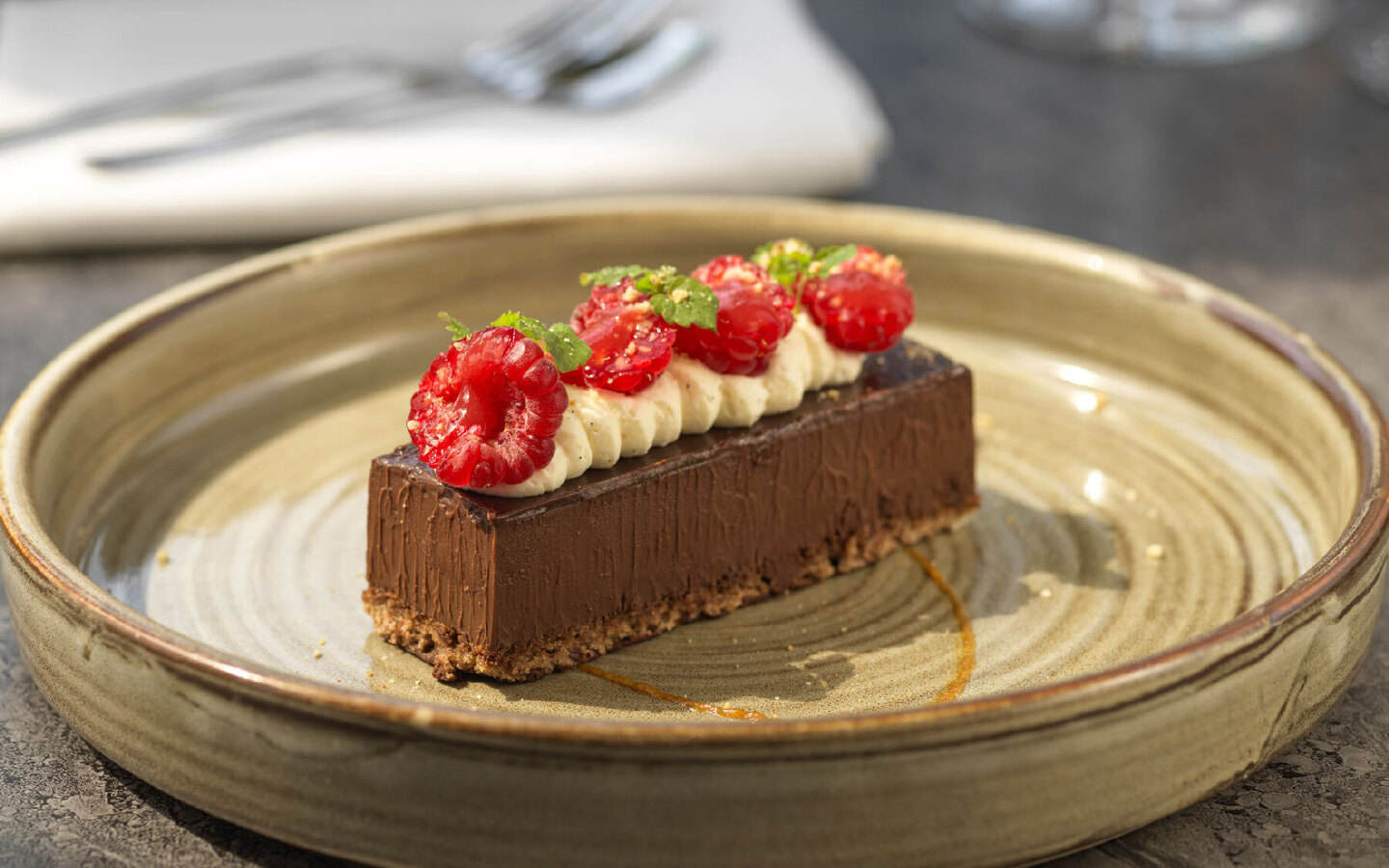 Decadent chocolate tart slice topped with piped cream, fresh raspberries and mint on a rustic ceramic plate at a restaurant table.