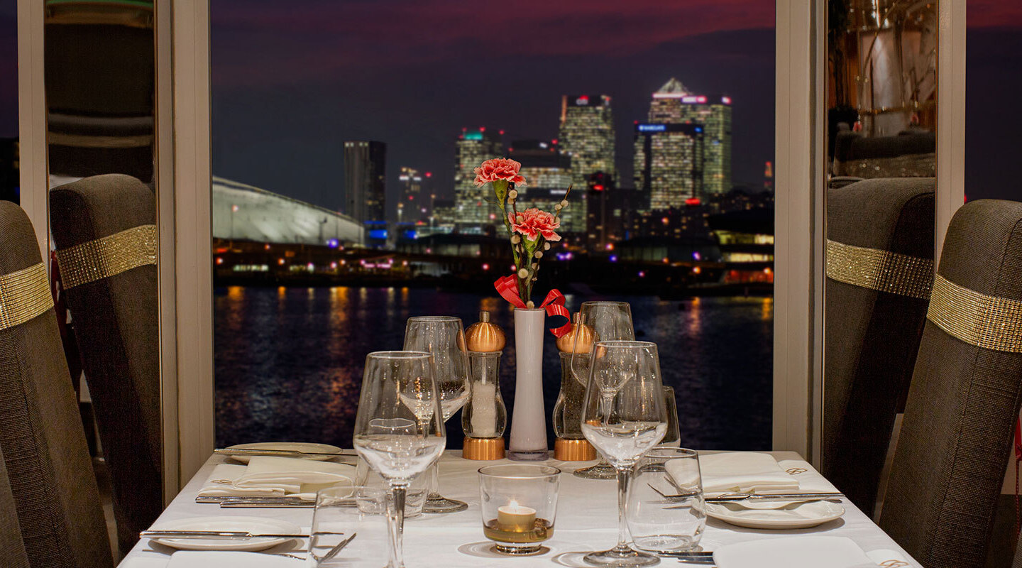 Candlelit waterfront table with white linen, crystal glassware, pink flowers and gold-banded high-back chairs, city skyline at night