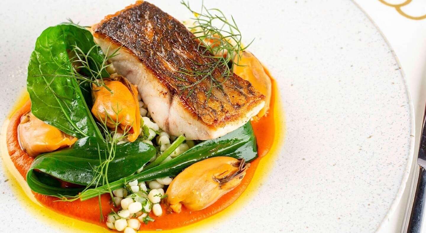 Seared fish fillet with mussels, pearl couscous, wilted greens and red pepper sauce, plated for hotel fine-dining.