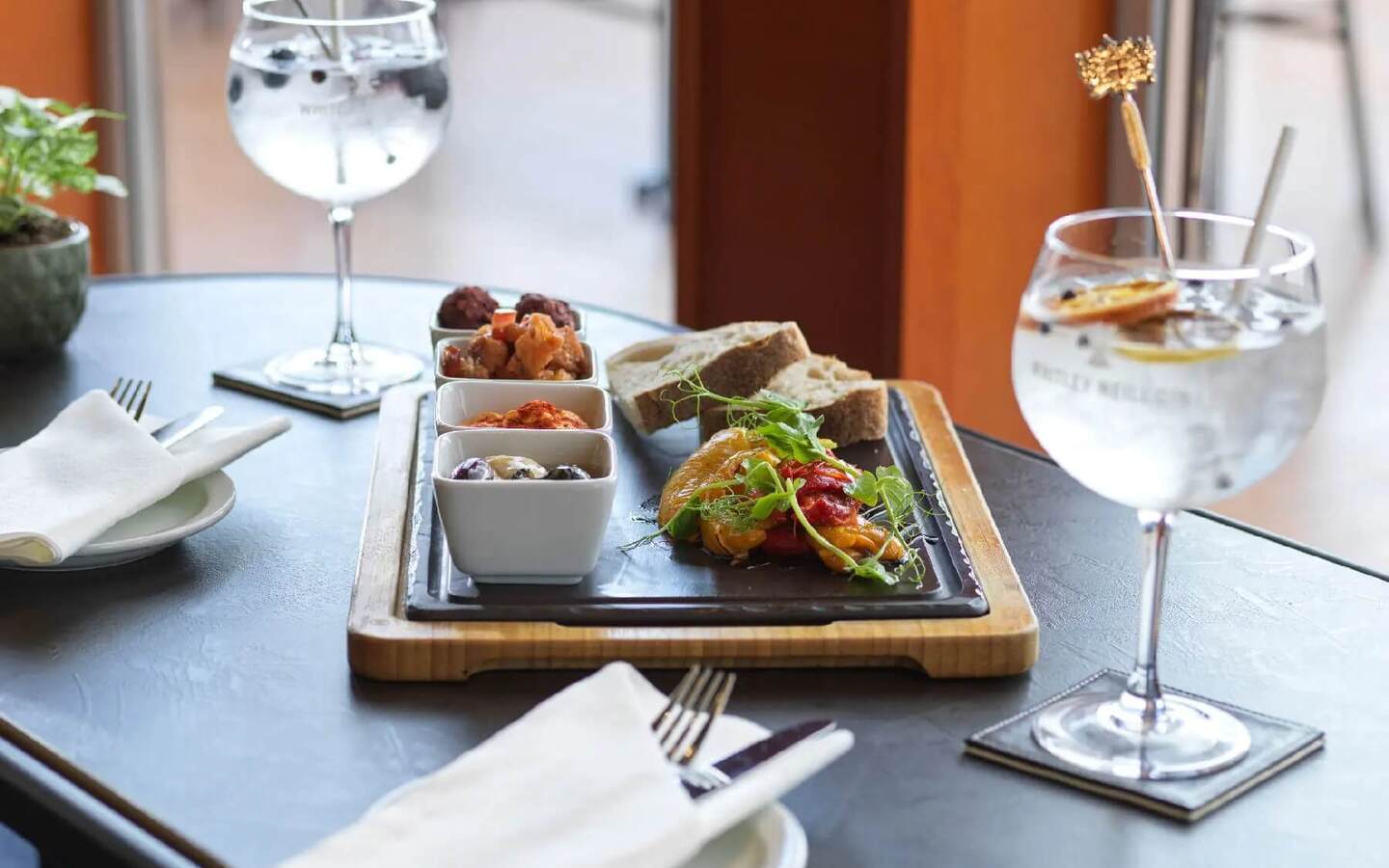Tapas sharing board with olives, dips, roasted peppers and bread on a wooden tray, served with two gin-and-tonic cocktails.