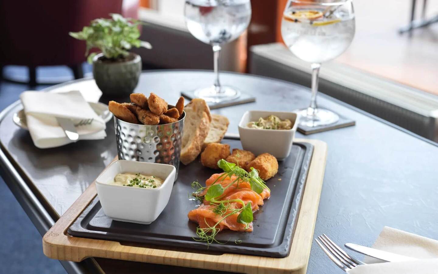 Smoked salmon and fried bites on slate board with dips, crusty bread and two gin-and-tonic glasses at hotel bar.