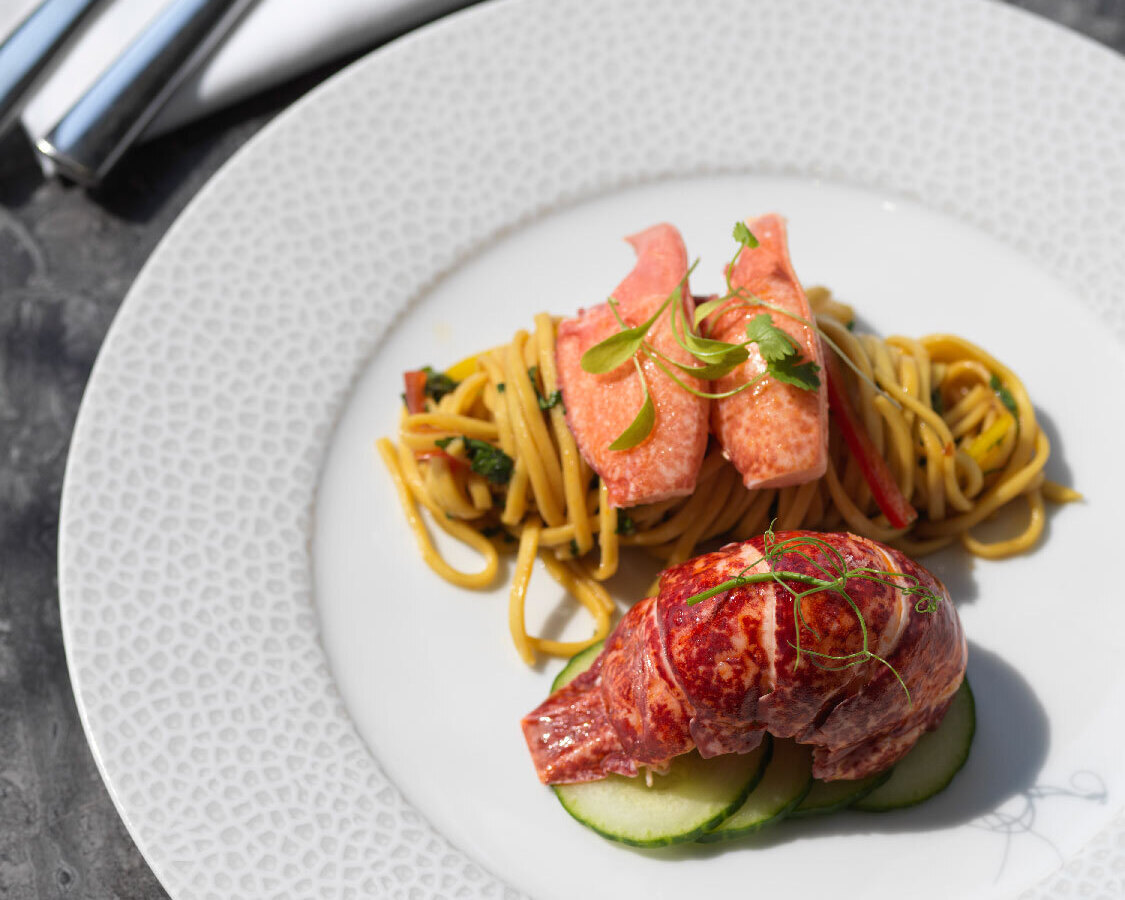 Plated lobster tail with tossed noodles, cucumber slices and microgreens on a textured white dinner plate, fork and knife.