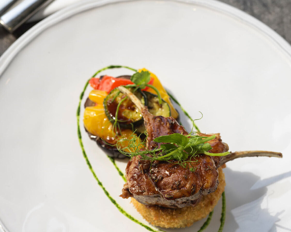 Seared lamb chops on a crispy potato cake with roasted cherry tomatoes and zucchini, herb oil swirl on white plate