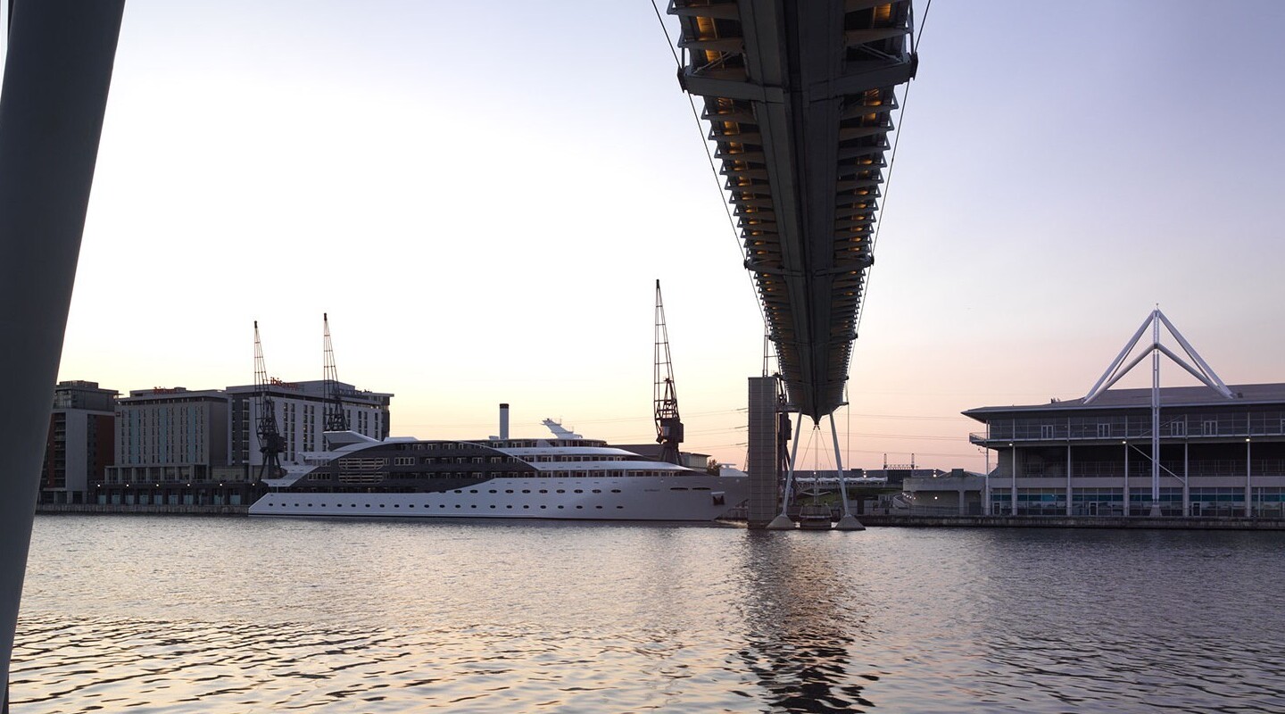 Luxury yacht moored beside modern waterfront hotels and a riverside conference center under an elevated pedestrian bridge at dusk.