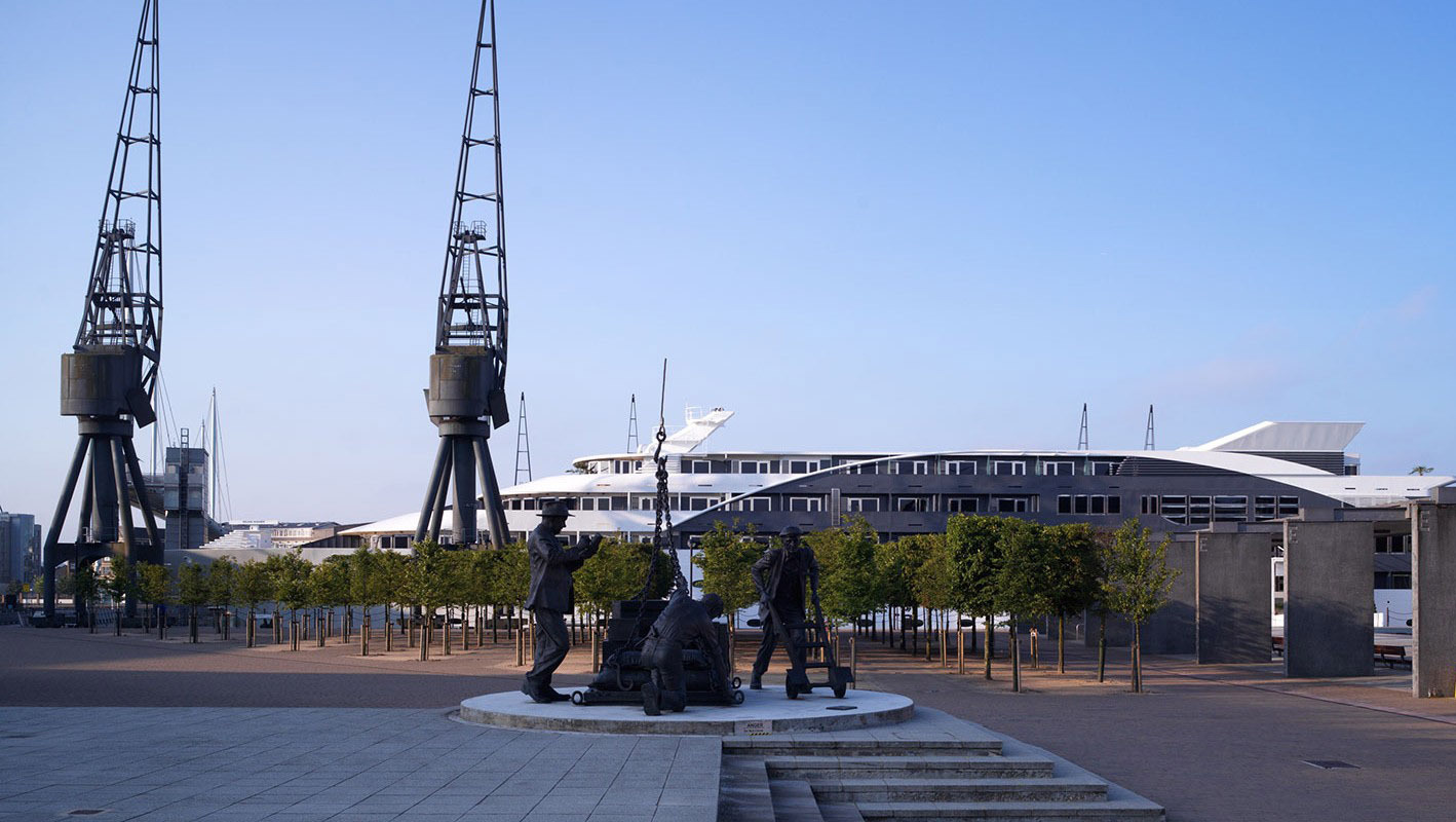 Waterfront promenade with tree-lined walkway, bronze dockworker statue, moored white superyacht and tall industrial cranes.