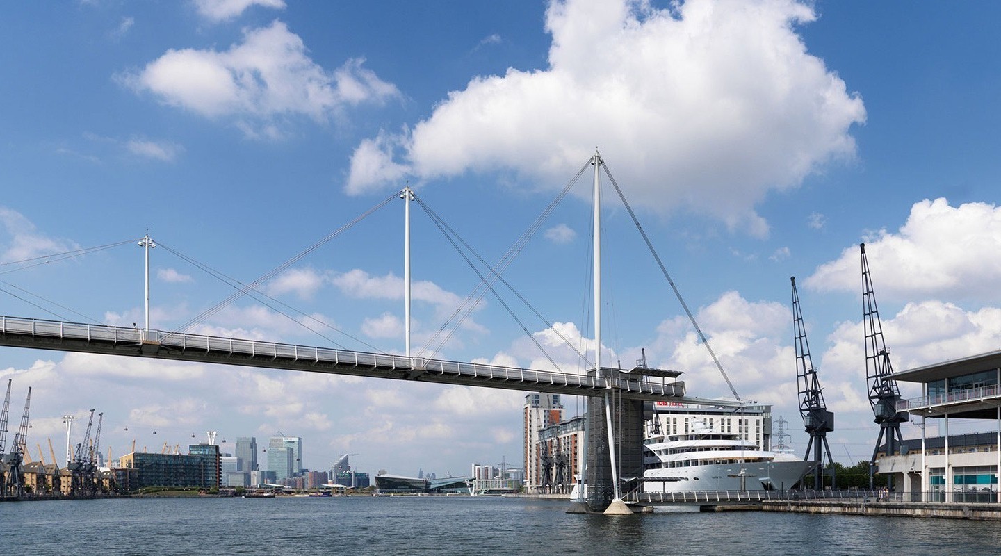 Riverside promenade and modern cable-stayed footbridge with a docked luxury yacht, historic cranes and city skyline views.