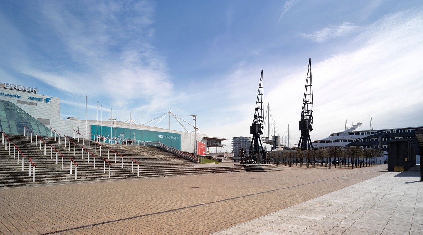 Waterfront ExCeL London convention center with broad stone steps and glass atrium, dockside cranes and moored yachts on a sunny plaza