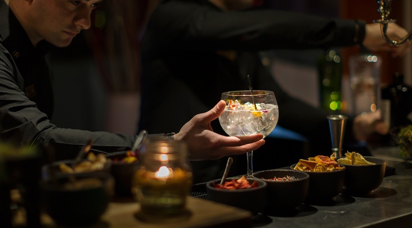 Bartender in black serving a large iced cocktail with citrus garnish at a candlelit hotel bar with bowls of snacks.