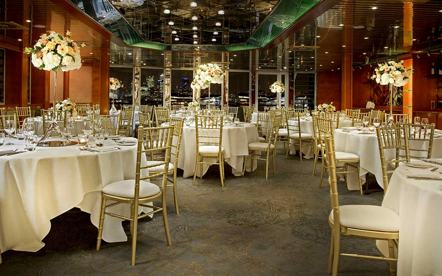 Elegant banquet room with white-linen tables, gold chairs, mirrored ceiling and tall floral centerpieces with city skyline view
