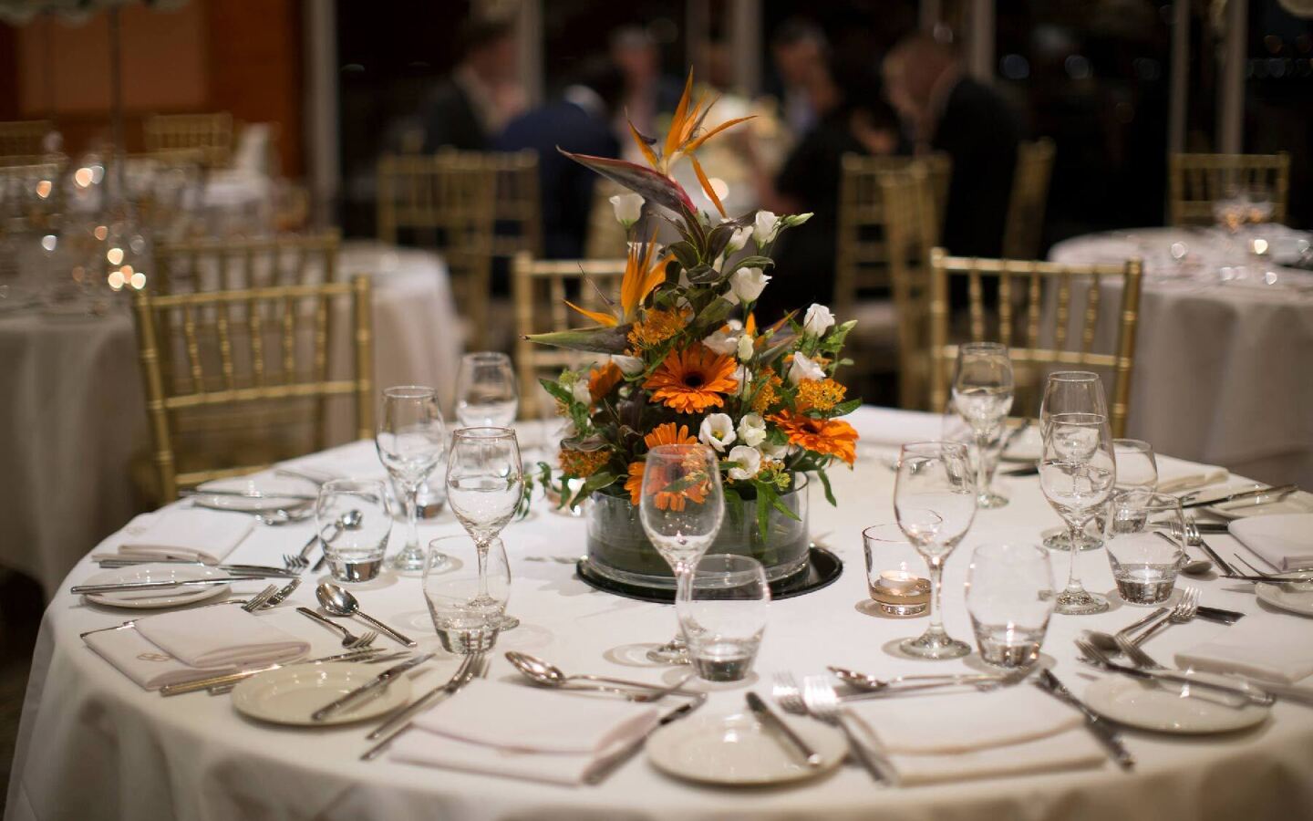 Round table with white linen, formal place settings, orange floral centerpiece and soft candlelight, gold Chiavari chairs