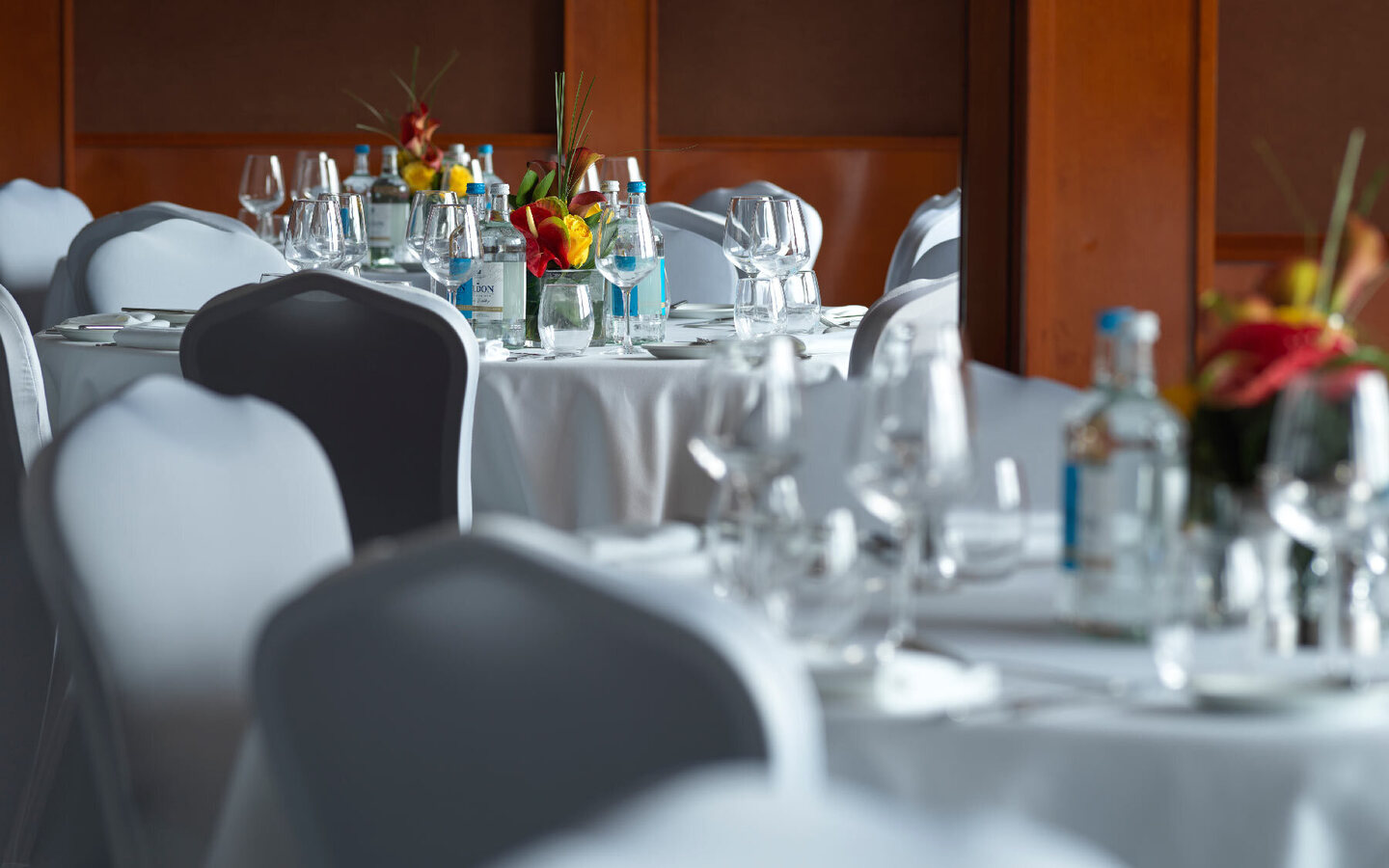 Elegant banquet tables with white linens and covered chairs, glassware, bottled water and tropical floral centerpieces.