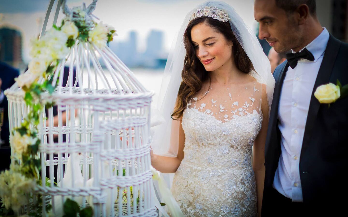 Bride in lace gown with jeweled veil and groom in tux beside a flower-adorned white birdcage at a waterfront wedding venue.