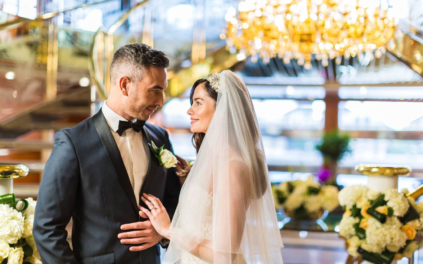 Bride and groom smile in hotel lobby beneath a crystal chandelier, surrounded by wedding flowers and a sweeping staircase