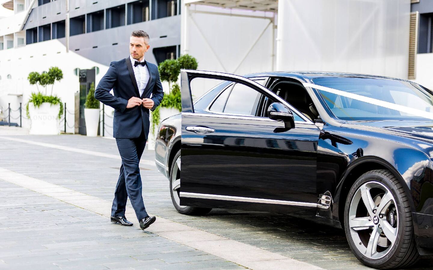 Doorman in tuxedo beside a black luxury car at a hotel entrance, adjusting his jacket as the car door stands open.