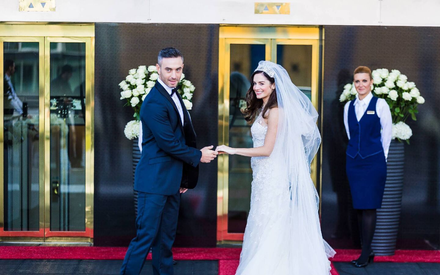 Newlyweds holding hands on a red carpet at a grand hotel entrance with gold doors, white rose displays and a concierge.
