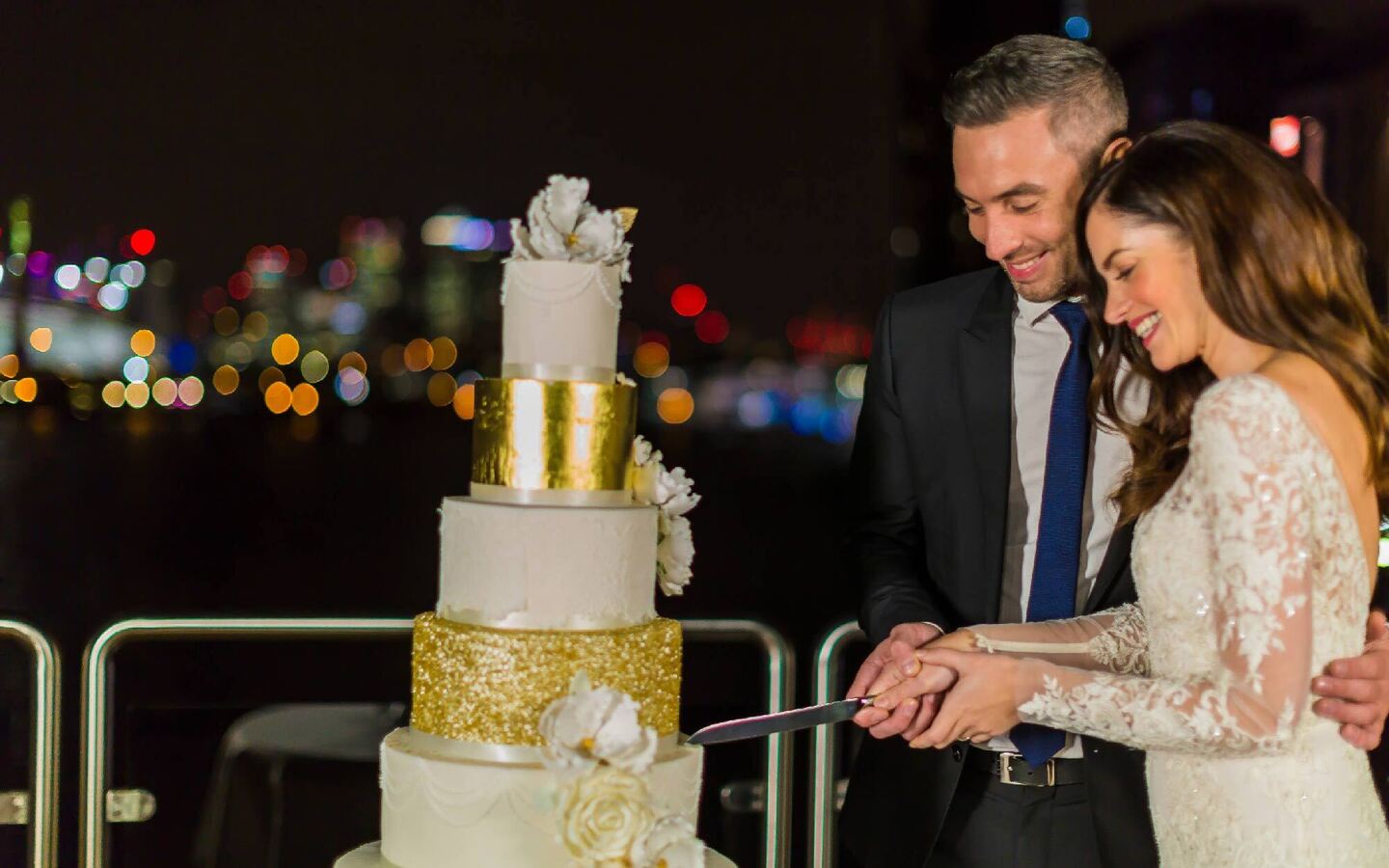 Newlyweds in lace gown and suit cut a multi-tier white-and-gold wedding cake on a waterfront terrace with city lights.