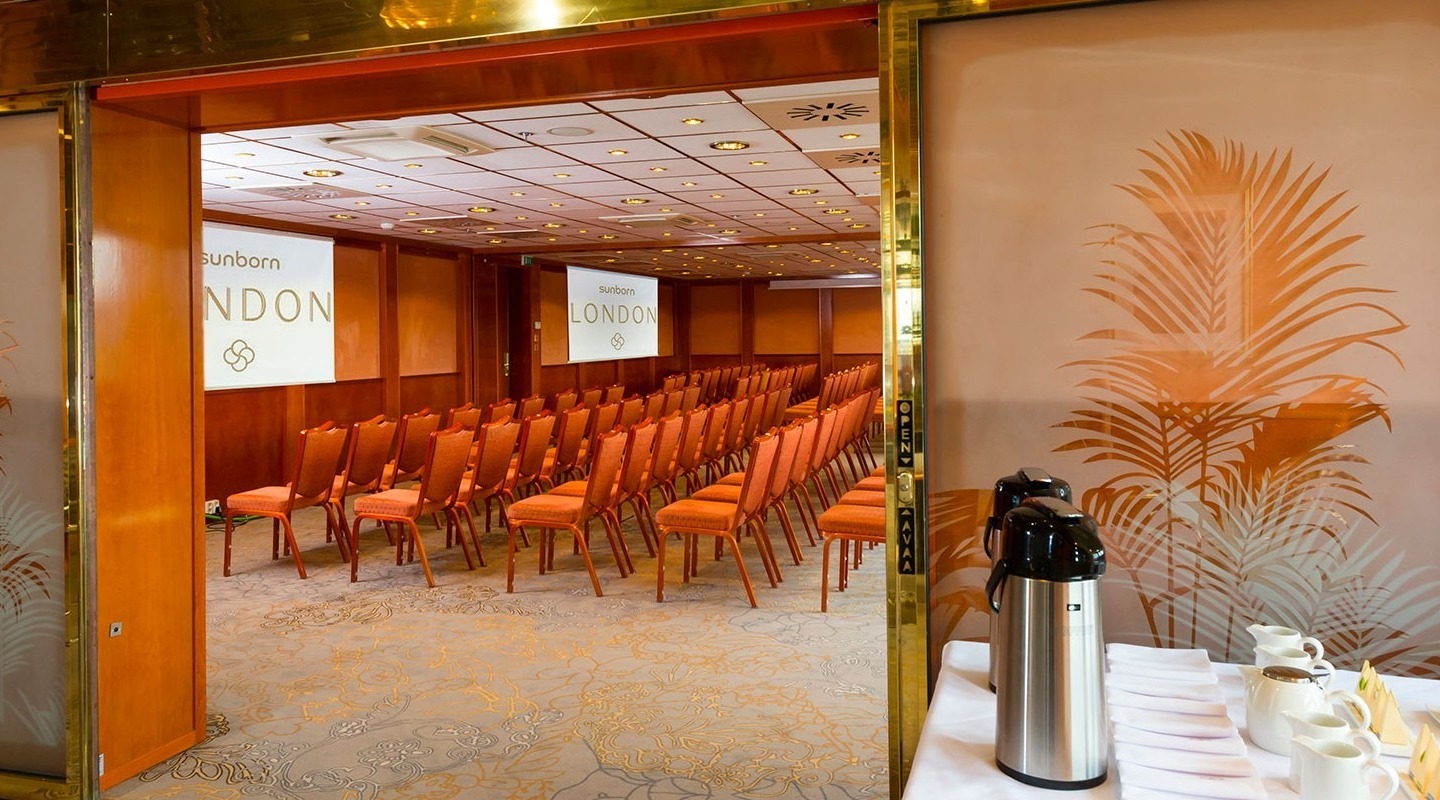 Theatre-style meeting room with rows of orange chairs, frosted glass palm doors and a self-serve coffee station with urns.
