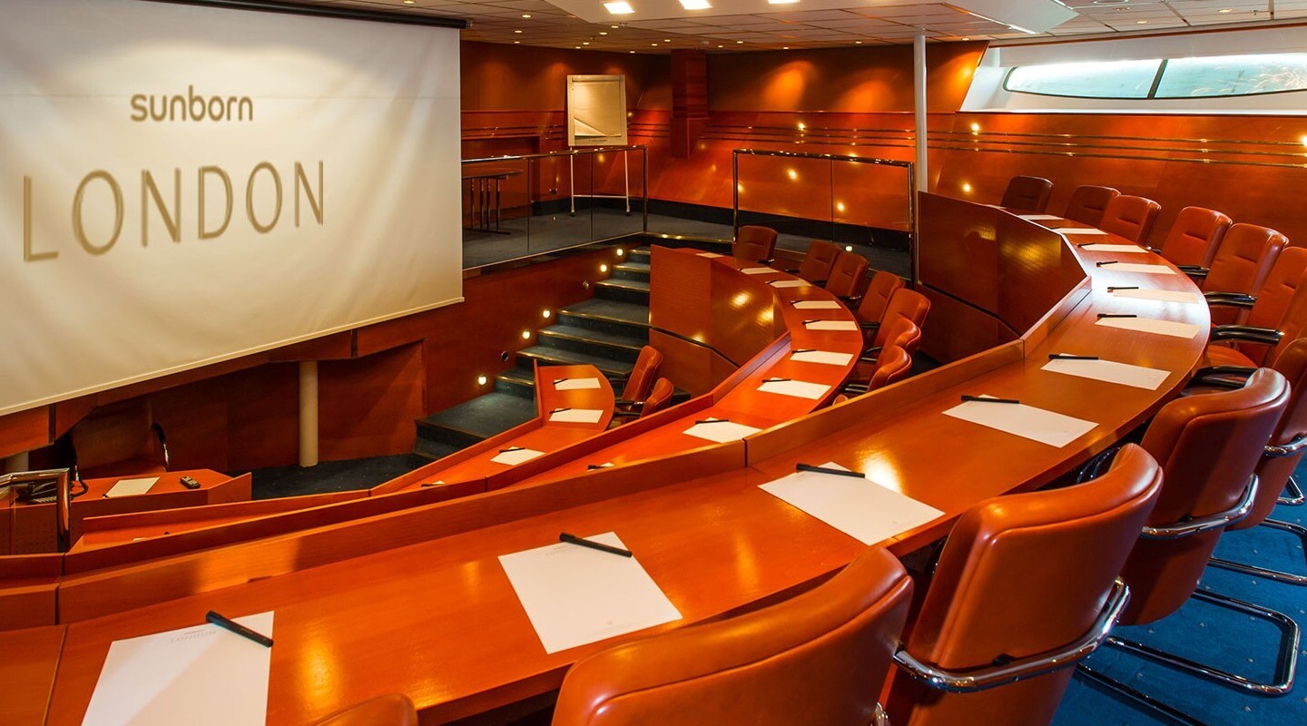 Tiered curved wood conference room with leather chairs, notepads and pens, warm lighting and large Sunborn London projection screen