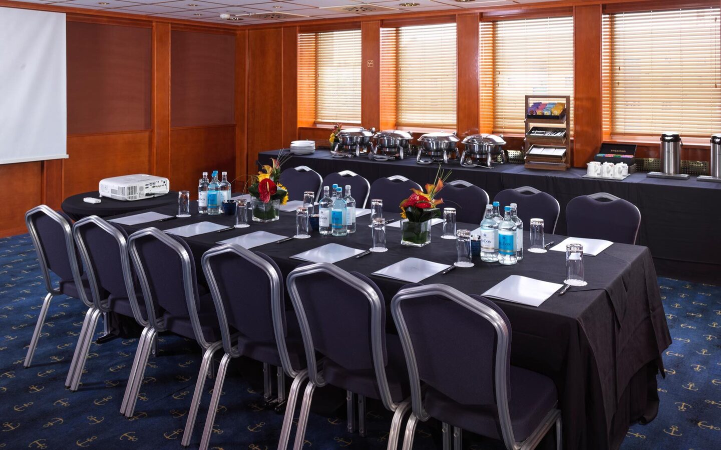 Conference room with black tablecloth, chairs, bottled water, flower arrangements, projector and buffet with coffee.