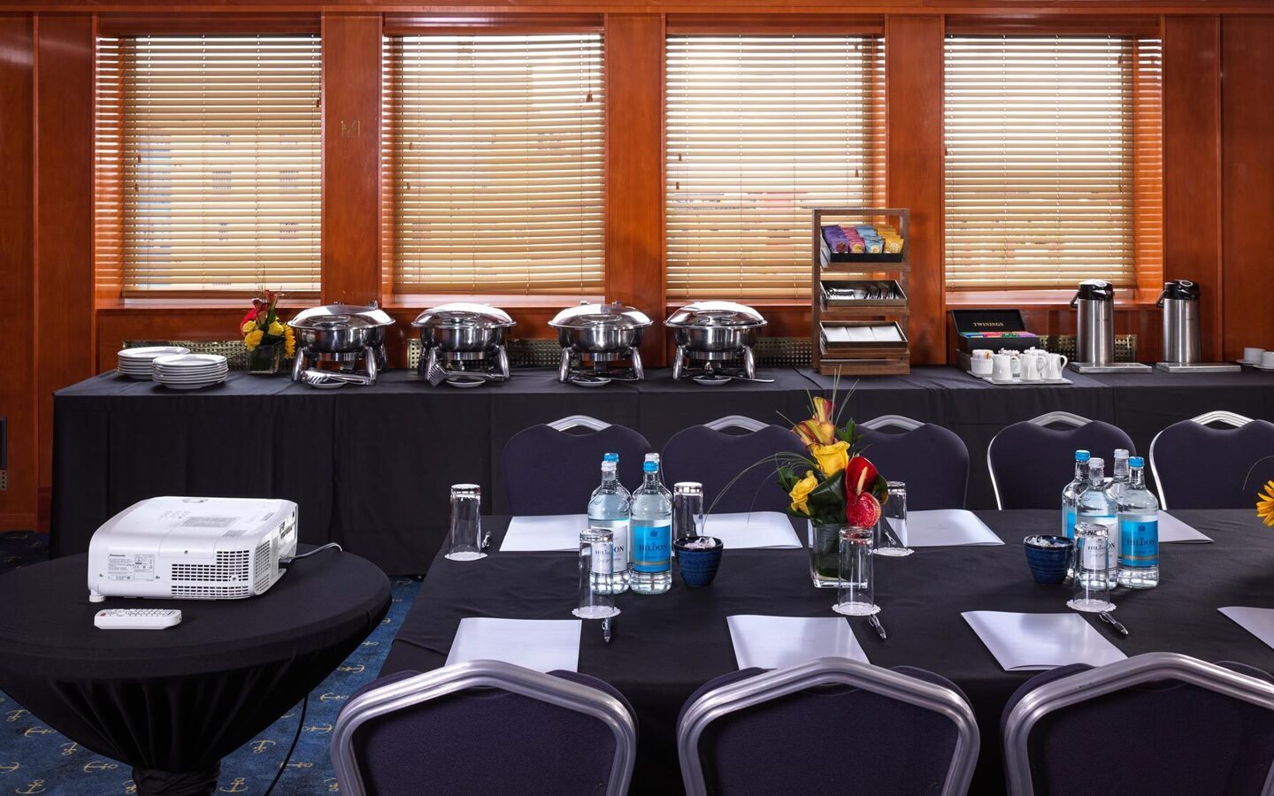 Conference room with projector, black-linen tables, bottled water, floral centerpieces, buffet chafing dishes and stainless coffee urns