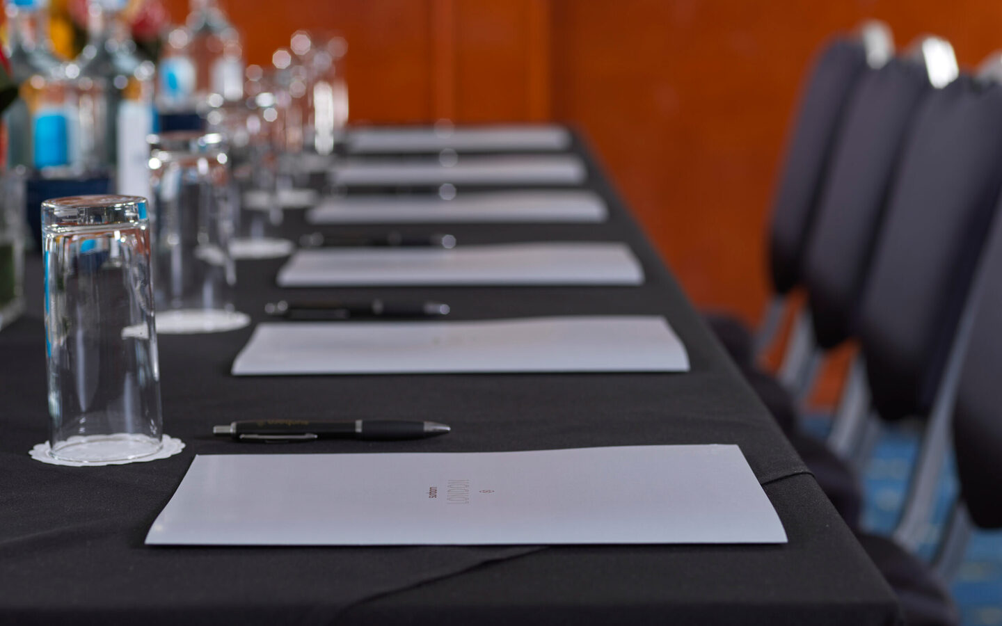 Hotel conference table with black tablecloth, notepads, pens, upside-down water glasses, bottled water and chairs.