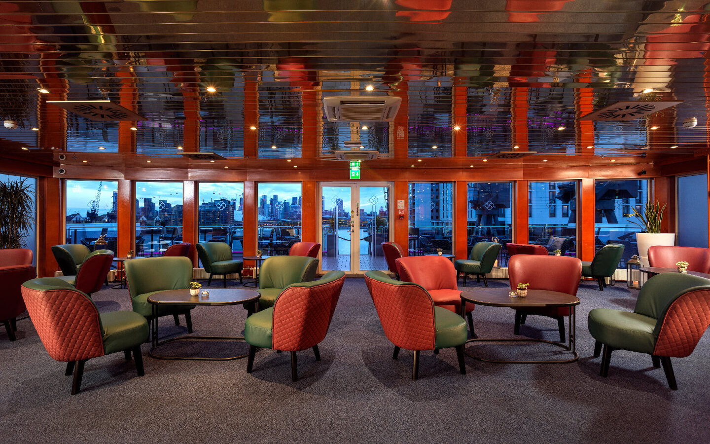 Hotel lounge with red and green armchairs, low tables and glossy wood ceiling, floor-to-ceiling windows with waterfront city and terrace view.