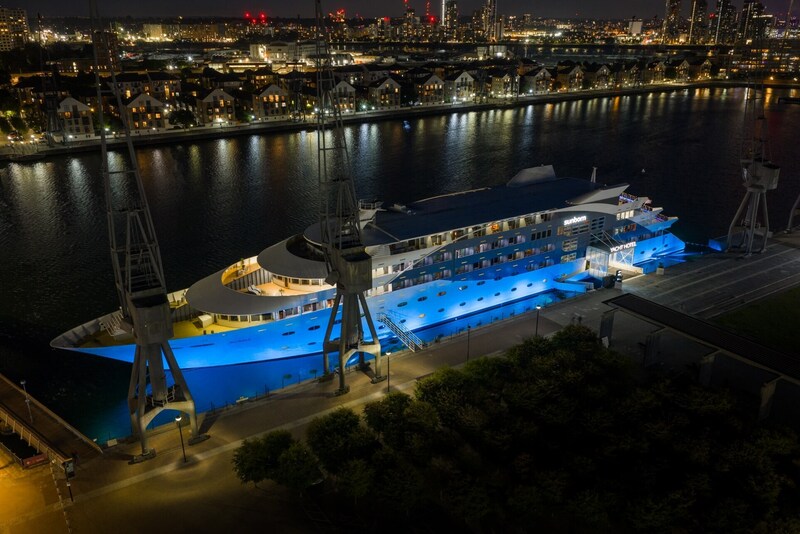 Floating yacht hotel lit in blue, docked at a riverfront promenade with city skyline lights reflected in the water.