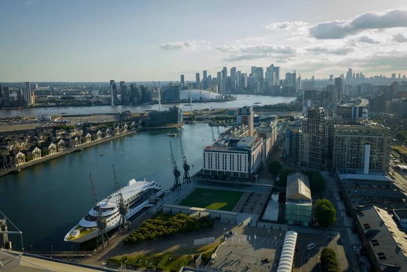 Riverside hotel view overlooking a marina with a moored yacht, Thames, O2 arena and Canary Wharf skyline at golden hour.