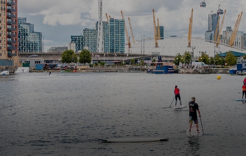Guests paddleboarding on a calm urban waterfront with modern apartments, cable cars and the O2 arena skyline in view