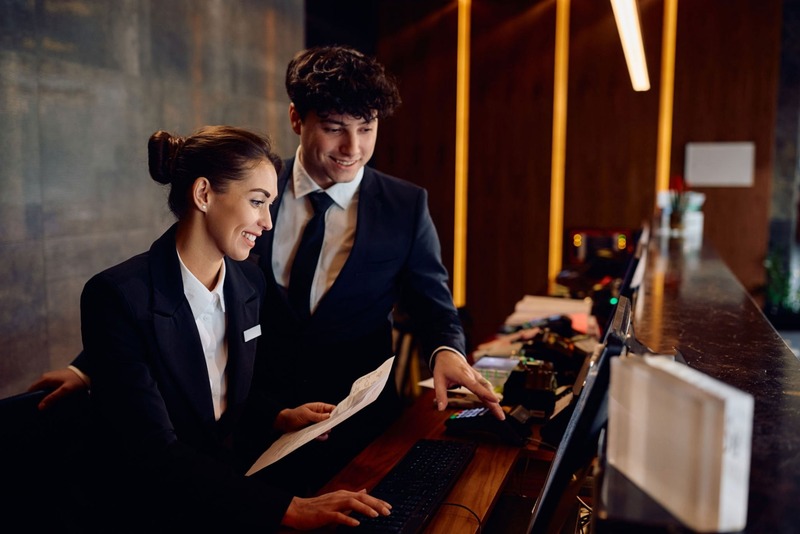 Two smiling front-desk staff in suits reviewing a guest folio at a modern reception with warm wood and pendant lights