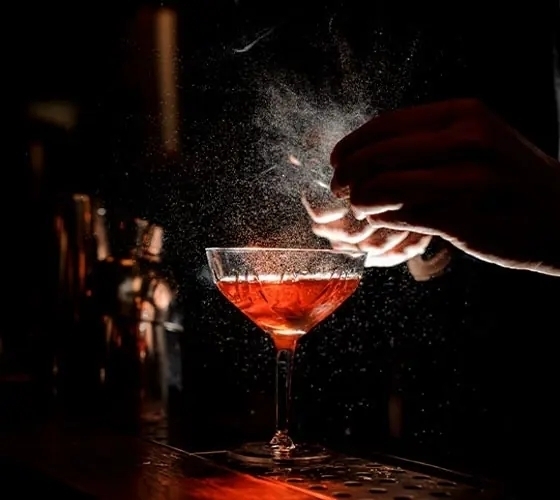 Warm-lit bar coupe with amber cocktail as bartender sprays citrus mist, showcasing a handcrafted drink and intimate speakeasy ambiance.