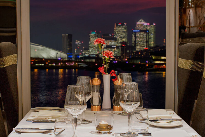 Candlelit waterfront table with white linen, crystal glassware, pink flowers and gold-banded high-back chairs, city skyline at night
