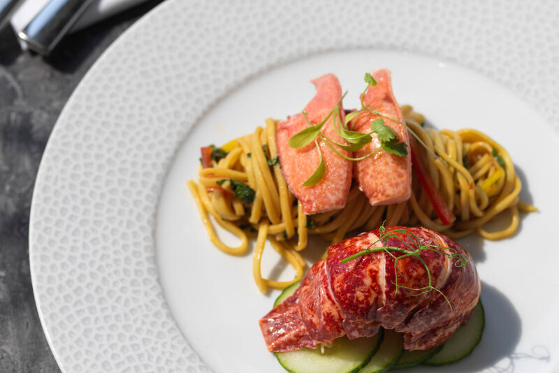 Plated lobster tail with tossed noodles, cucumber slices and microgreens on a textured white dinner plate, fork and knife.