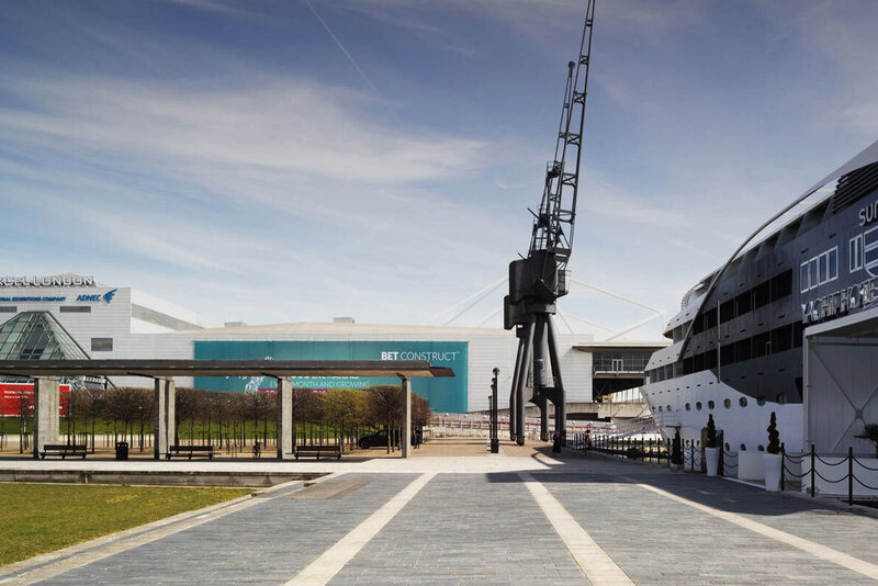 Luxury Sunborn yacht hotel moored beside ExCeL London, waterfront promenade with benches, lawn and a historic dock crane under a blue sky