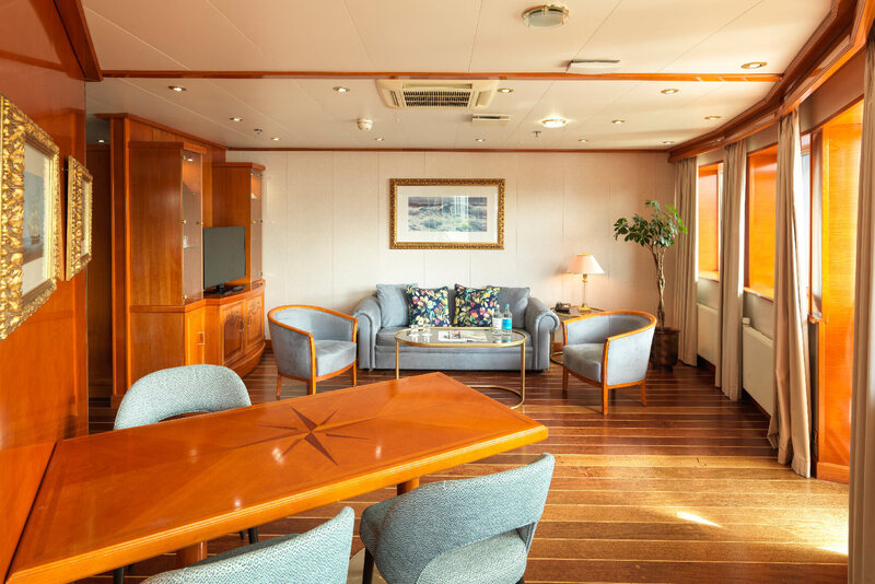 Luxury suite lounge with warm wood finishes, dining table, plush sofa and chairs, coffee table, lamp and sunlit windows.
