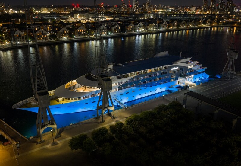 Floating yacht hotel lit in blue, docked at a riverfront promenade with city skyline lights reflected in the water.