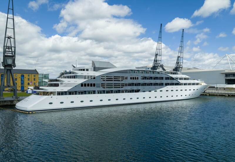 Large white luxury yacht docked at a waterfront terminal with multi-deck balconies, panoramic windows and tall dock cranes.