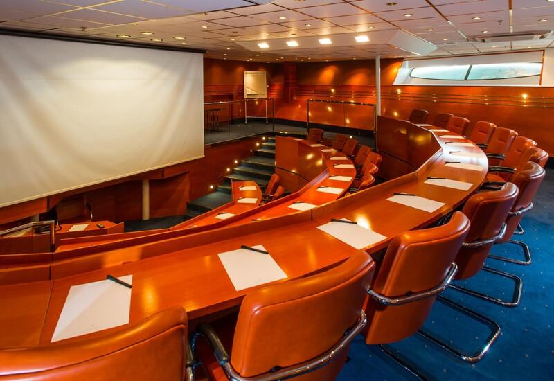 Tiered conference theater with large projection screen, curved wood desks, leather swivel chairs, pens and pads, soft lighting and narrow windows.