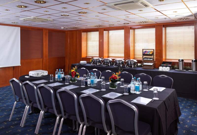 Hotel meeting room with conference tables set with notepads, bottled water, projector; buffet chafers, wooden blinds and recessed lights.