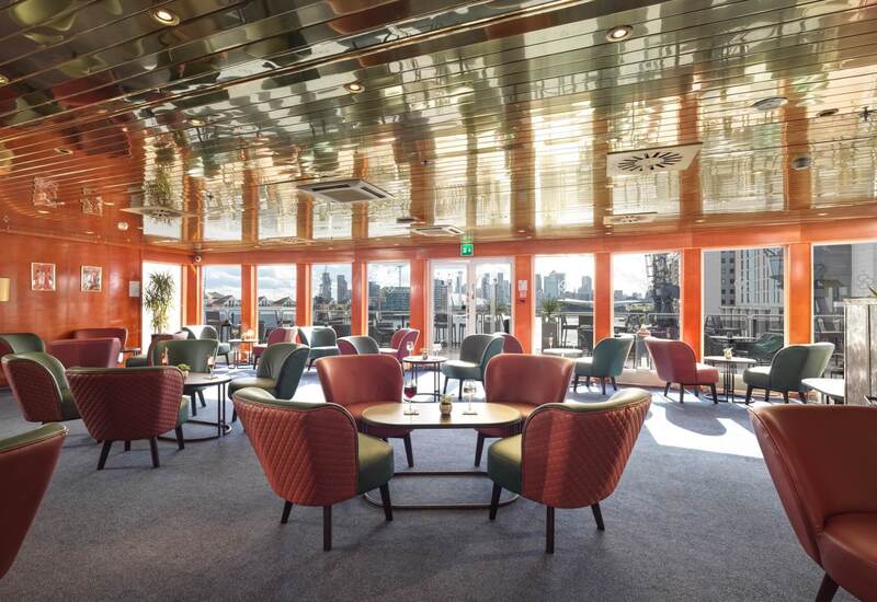 Sunlit hotel lounge with quilted armchairs, tables with drinks, reflective ceiling and city skyline terrace view