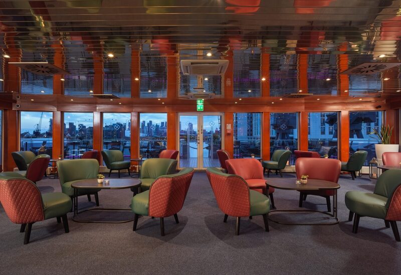 Hotel lounge with green and red quilted armchairs, low tables, reflective ceiling and glass doors to a waterfront terrace and city skyline.