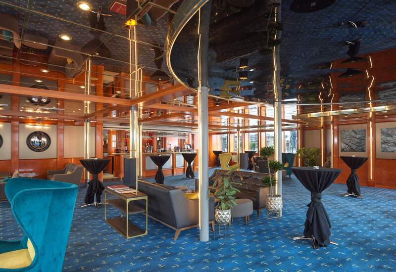 Nautical-themed lounge with mirrored ceiling, warm wood beams, bar, teal chairs and sofas, anchor-patterned blue carpet.