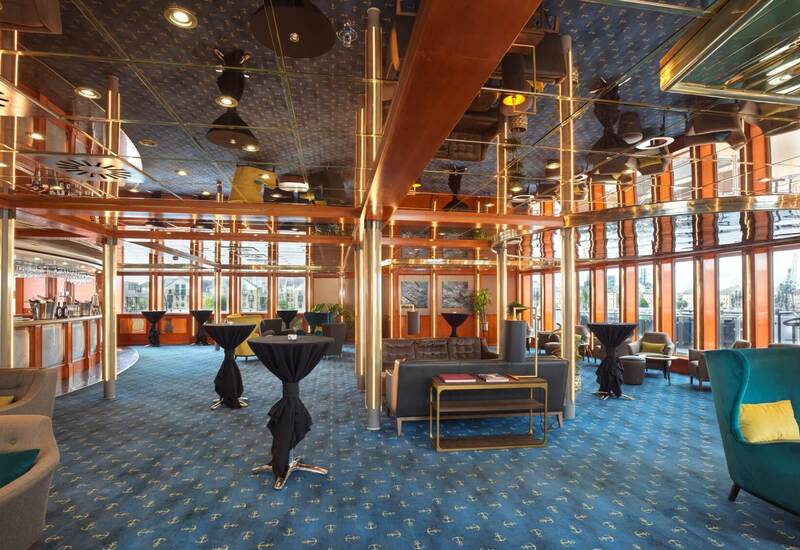 Waterfront hotel lounge with mirrored ceiling, brass and wood accents, anchor-pattern blue carpet, bar, cocktail tables and plush seating.