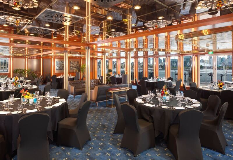 Elegant waterfront banquet room with round black-clothed tables, formal place settings, mirrored ceiling and city skyline views.