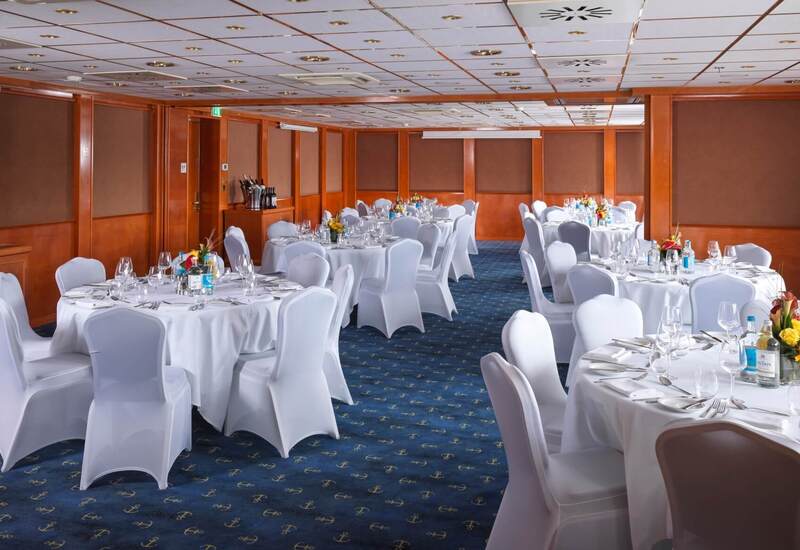 Hotel banquet room with round tables in white covers, floral centerpieces, glassware, bottled water and wood-paneled walls.