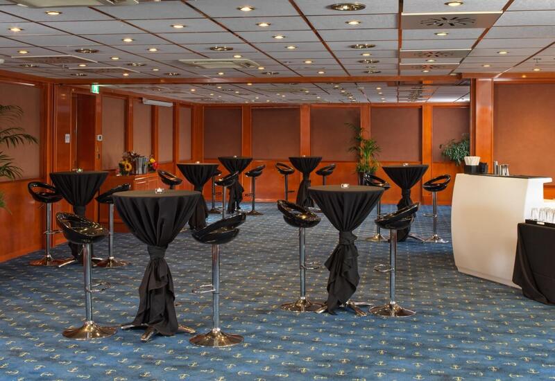 Wood-paneled hotel event room set for a cocktail reception with black-draped high tables, glossy bar stools, bar station and wine buckets.