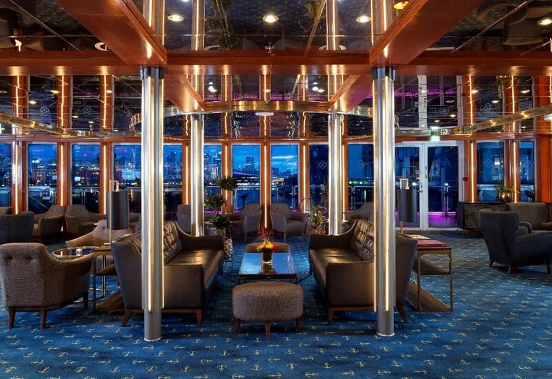 Upscale hotel lounge with leather sofas, armchairs, mirrored ceiling, metal-lit columns, anchor carpet and waterfront view.